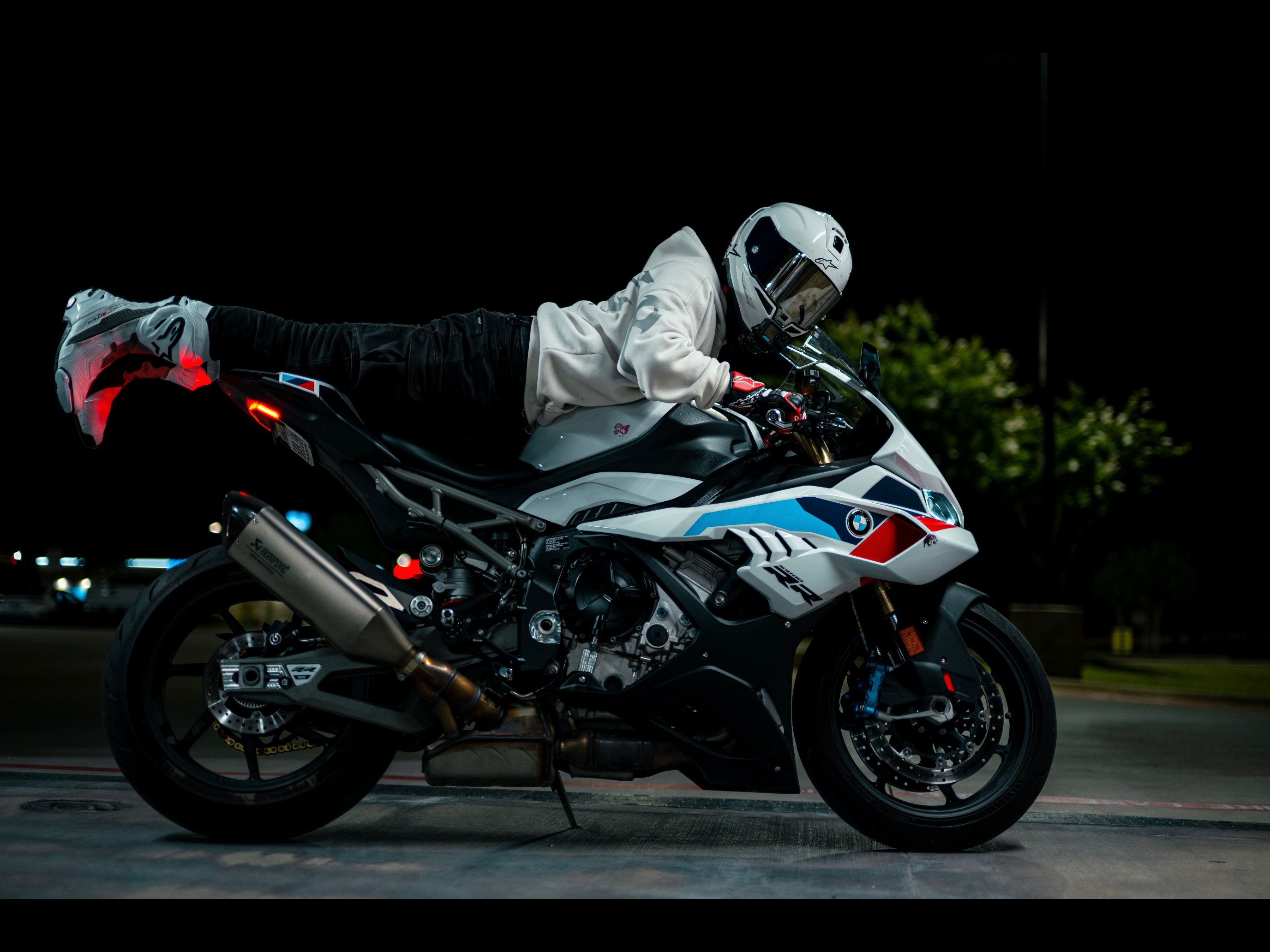 Blaze stretched flat along his BMW S1000RR in a superman pose at night, one boot kicked out behind him, parked lot lit only by the bike's headlight and city glow.