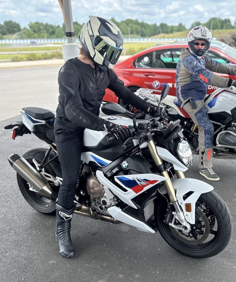 Blaze seated on a white BMW S1000R streetfighter motorcycle at a track day event, with another rider on a BMW nearby.